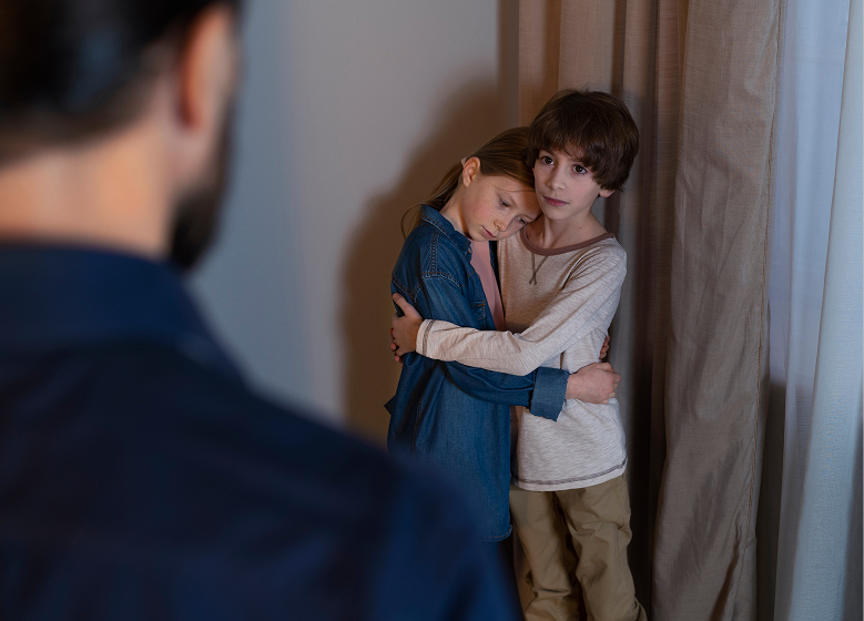 A boy and girl hug, standing by curtains, appearing scared or worried. A person's back is in the foreground, creating a tense atmosphere.