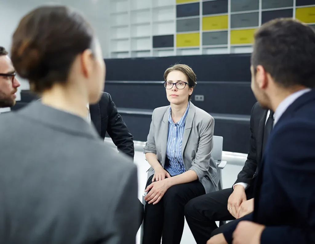 A woman in glasses sits attentively in a circle of seated professionals. She wears a gray blazer and looks engaged, set in a modern, minimalist office.