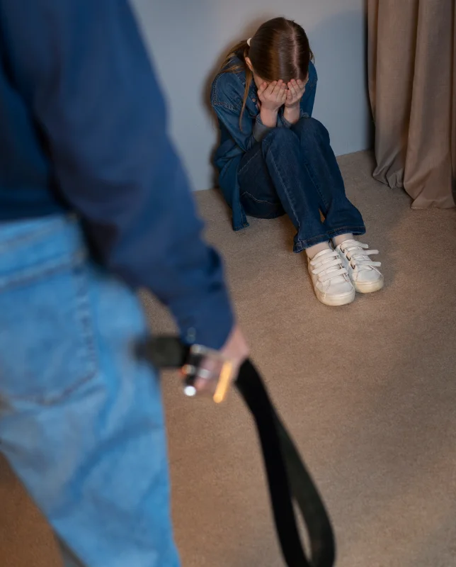 A child sits on the floor, covering her face with her hands, while an adult stands nearby, holding a belt.