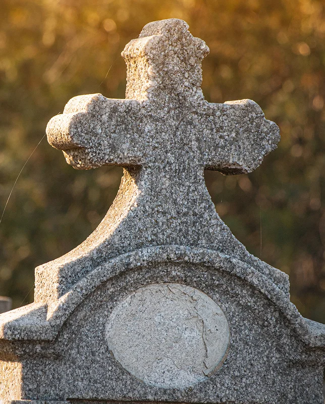 Stone grave cross with a weathered texture, illuminated by warm sunlight, against a blurred background of greenery. The image conveys a serene, timeless atmosphere.
