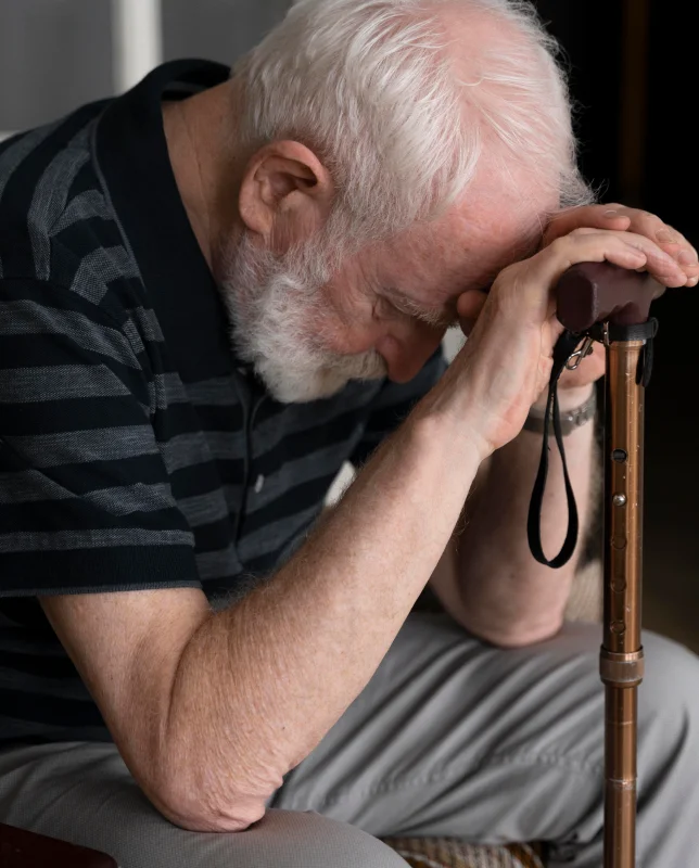 Elderly man with white hair leans on a cane, head bowed and hands clasped, wearing a striped shirt. The mood is reflective and somber.