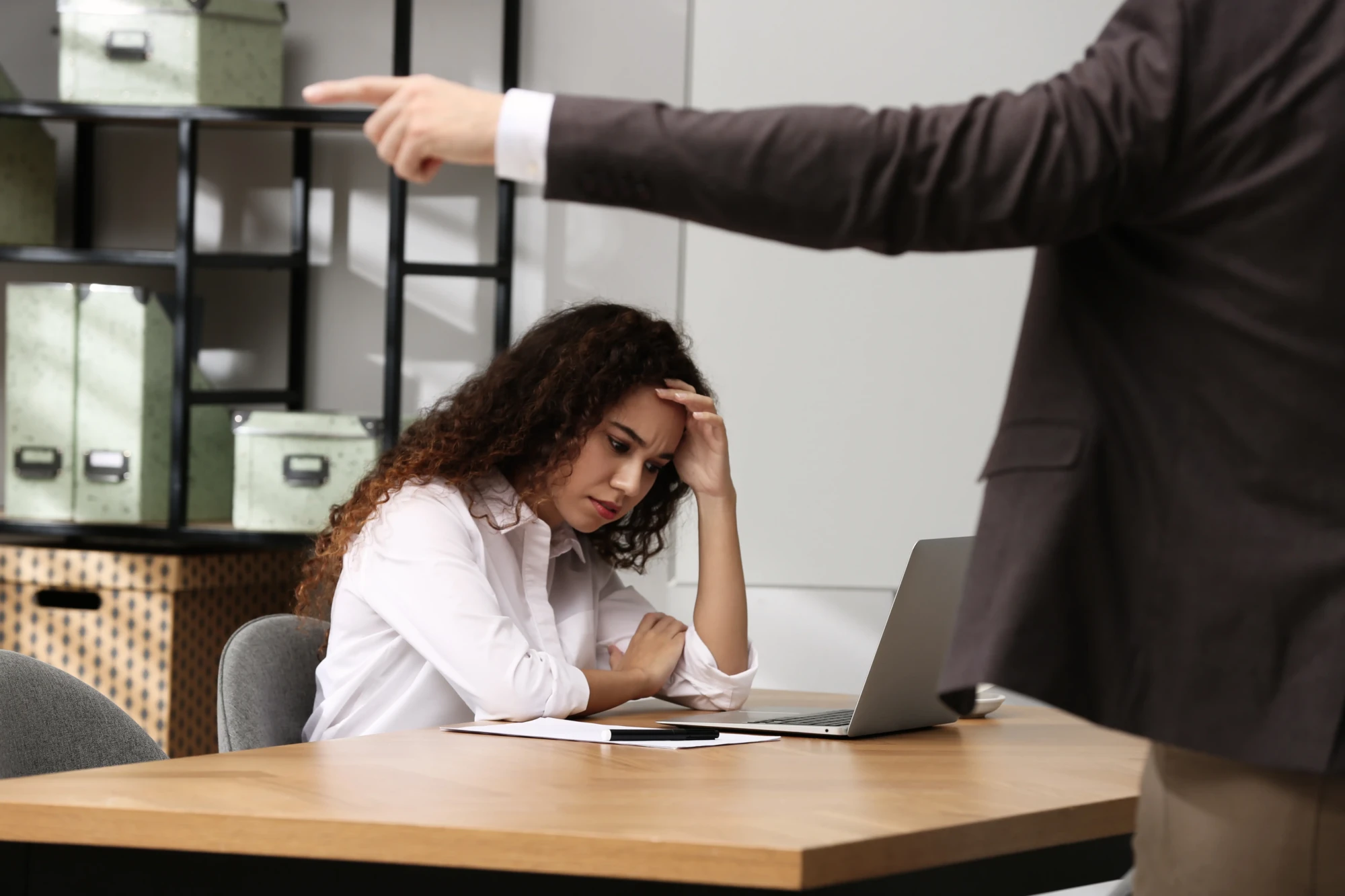An office scene shows a woman in a white blouse looking stressed at a desk with a laptop. A person in a suit points sternly at her, conveying tension.