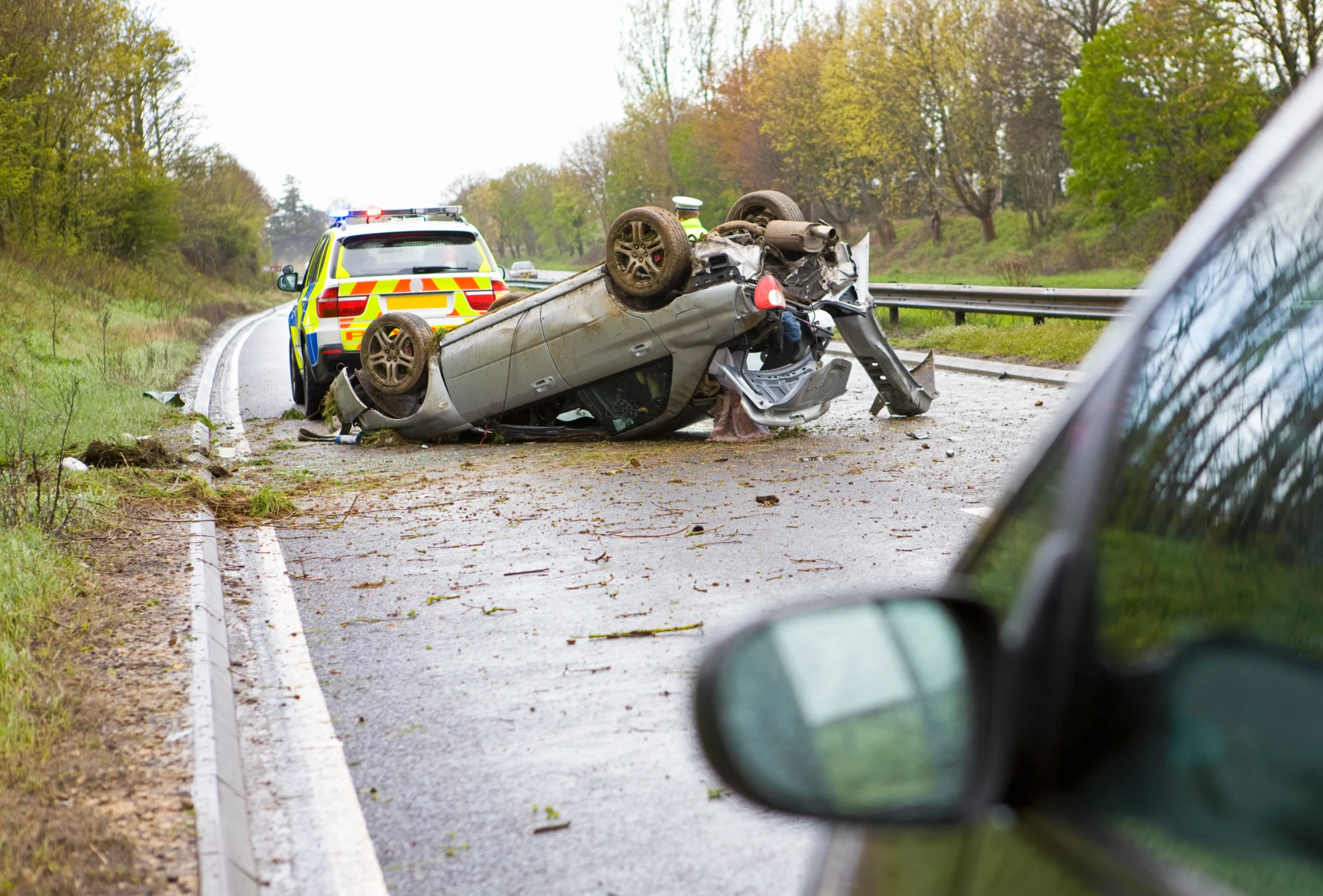 Overturned car on a wet road with police vehicle nearby. Trees line the road, creating a tense, urgent scene of a recent accident.