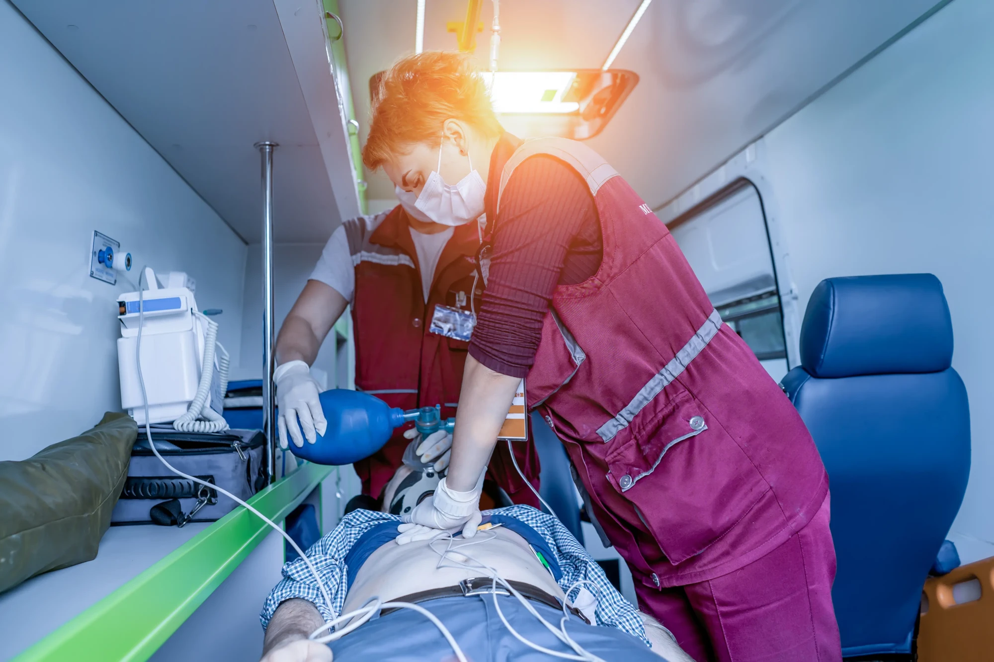 Two paramedics in red uniforms perform CPR on a patient in an ambulance. One uses a bag valve mask. The scene is urgent and professional.