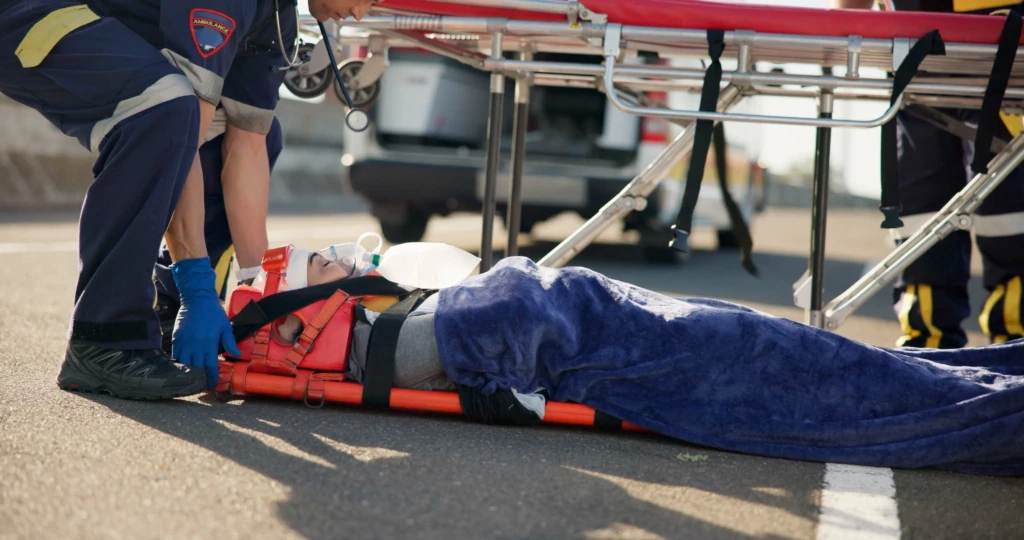 Paramedics tend to an injured person on a stretcher, wrapped in a blue blanket, on a road. The scene conveys urgency and care.