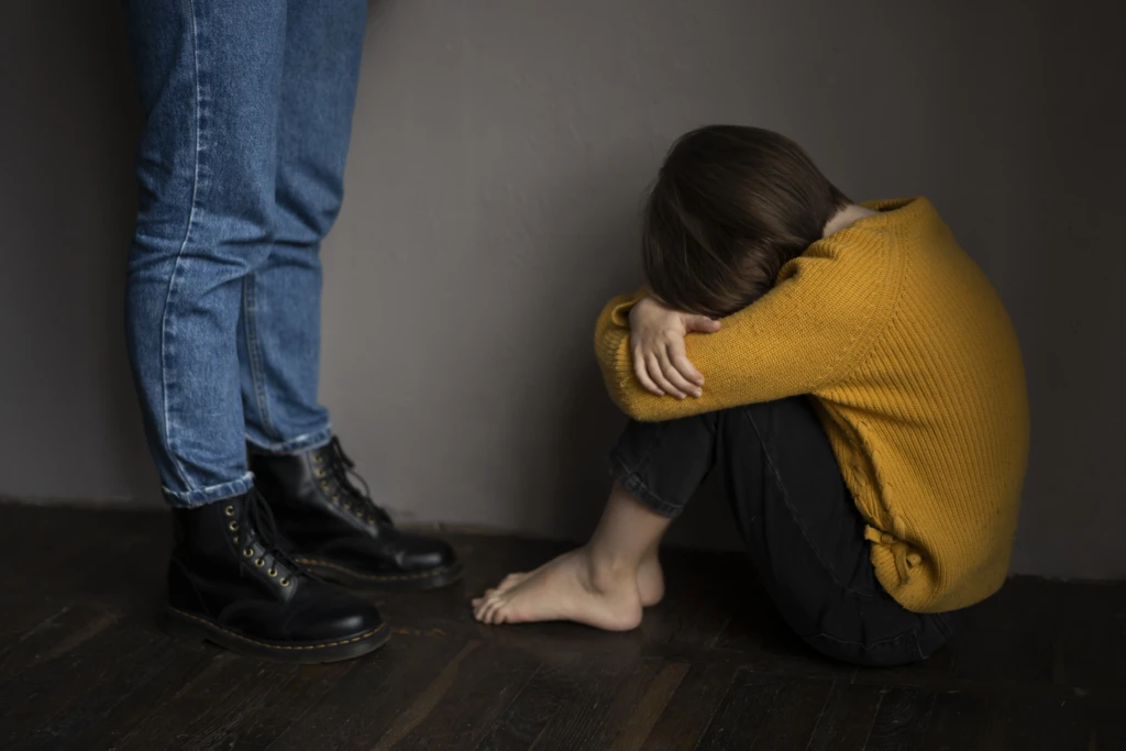 A child in a yellow sweater sits curled up on the floor with head resting on arms, conveying sadness. Nearby, an adult in jeans and boots stands.