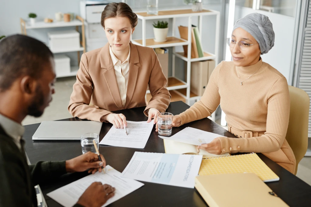 Three people in a meeting, sitting at a table with documents and laptops. The mood is focused and professional, with a shelf in the background.