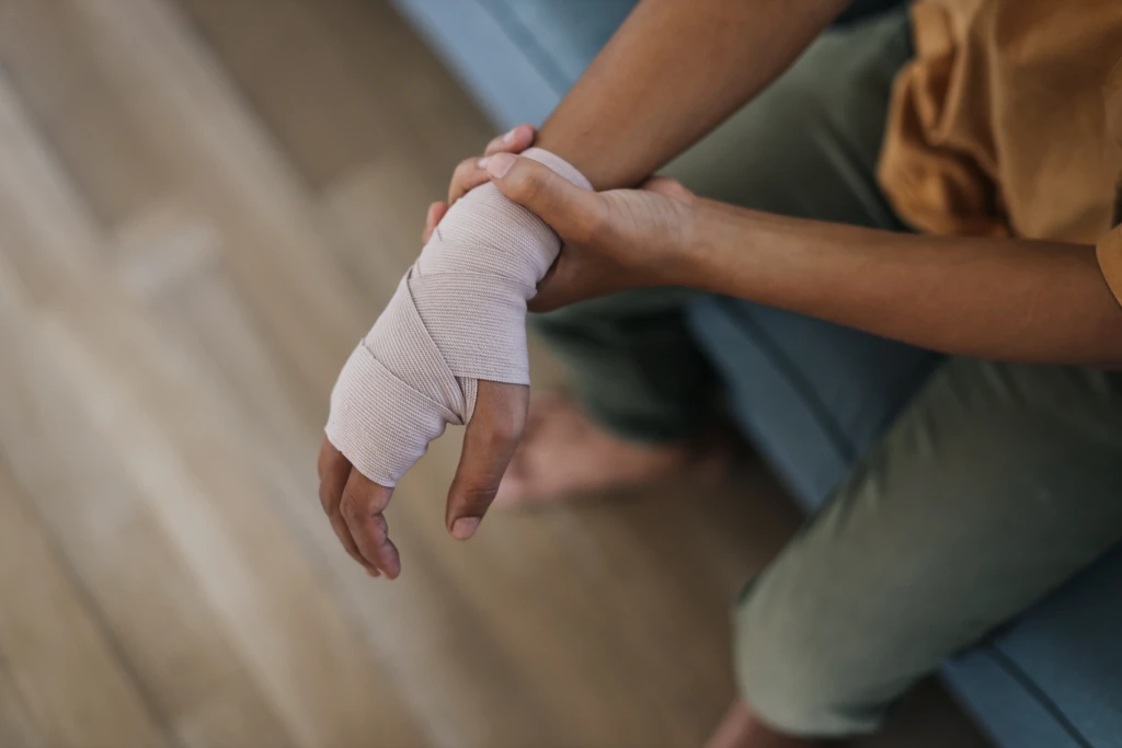 A person with a bandaged hand sits on a couch, gently holding their wrist, suggesting an injury. The image conveys a tone of care and recovery.