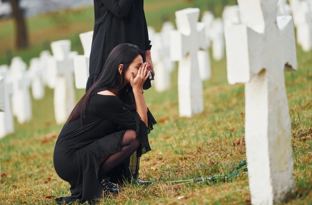 A woman in black kneels, covering her face, next to white crosses in a cemetery. The scene is somber, reflecting mourning and remembrance.