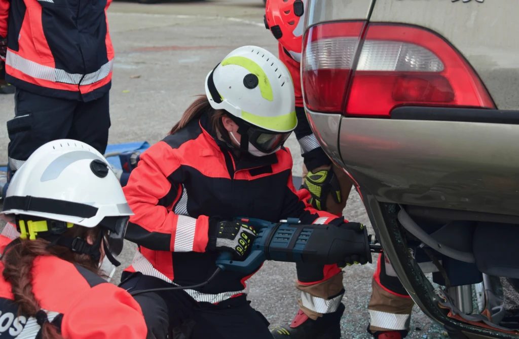 Rescue workers in helmets and protective gear use cutting tools on a car in a drill. The scene is focused, showing teamwork and urgency.