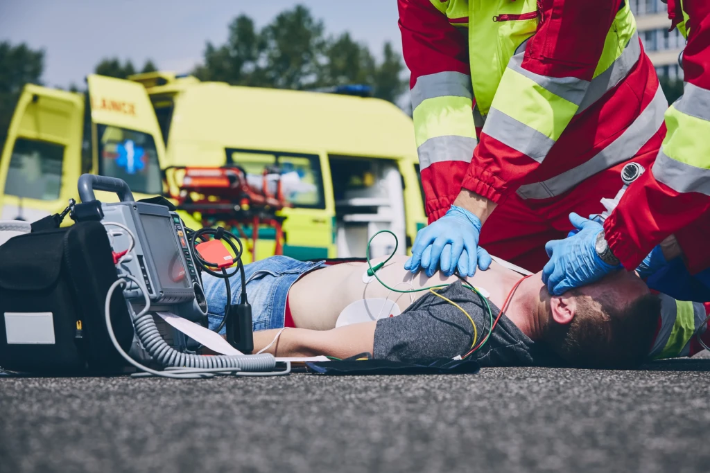 Paramedics perform CPR on a person lying on a road, connected to a defibrillator. An ambulance is parked in the background, conveying urgency.