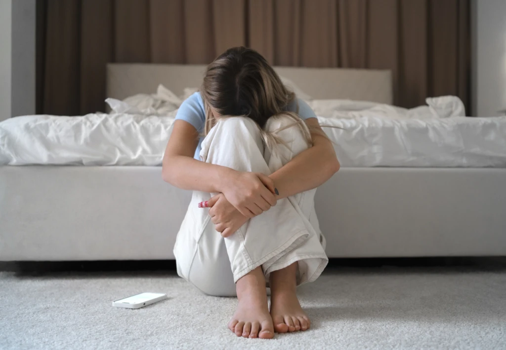 A person sits on the floor against a bed, head down on crossed arms, conveying sadness. A smartphone lies nearby on the carpet.