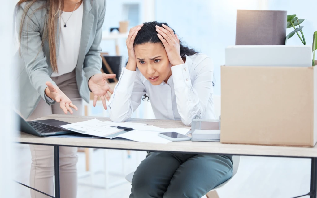 A stressed woman sits at a desk with her head in her hands, surrounded by papers and a laptop. Another person gestures in frustration beside her.