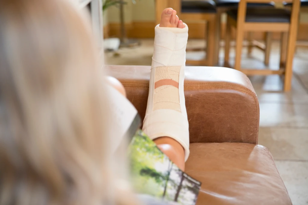 A person with a bandaged leg rests on a leather couch, reading a magazine. The room has a calm, cozy atmosphere, with wooden furniture in the background.