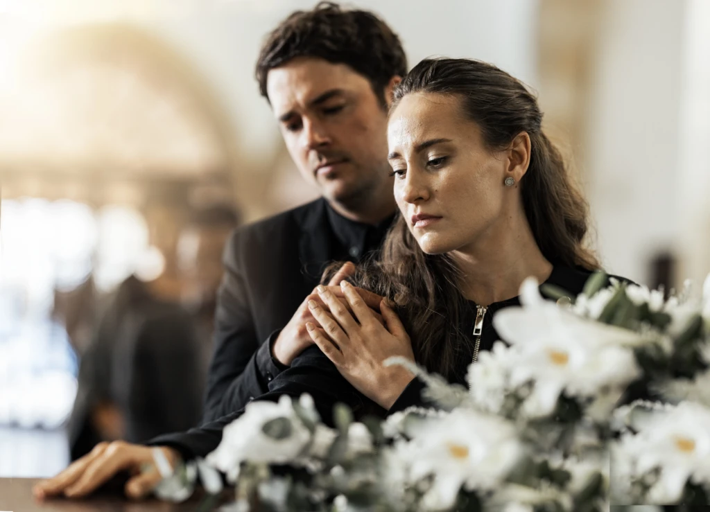 A man and woman in black attire stand close, appearing solemn in a bright room with blurred background. White flowers are in the foreground, in a funeral.