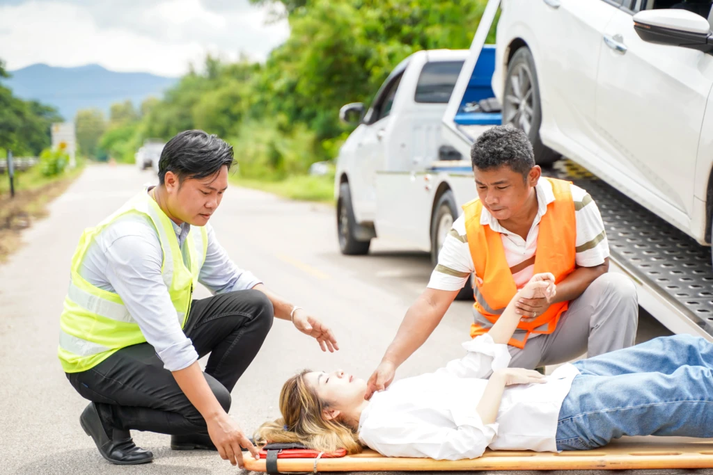 Two men in safety vests assist a woman lying on a stretcher by a road, near a white car and tow truck. The scene feels urgent and supportive.